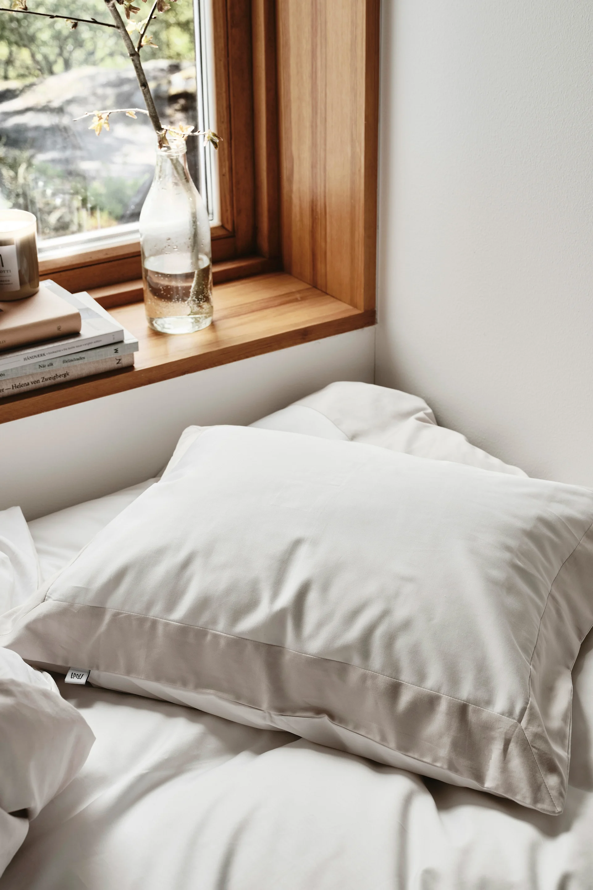 Vaaleanbeige tyyny valkoisella sängyllä, ikkunan vieressä, jossa on puinen ikkunalauta, kirjoja ja kasvinoksa pullossa.