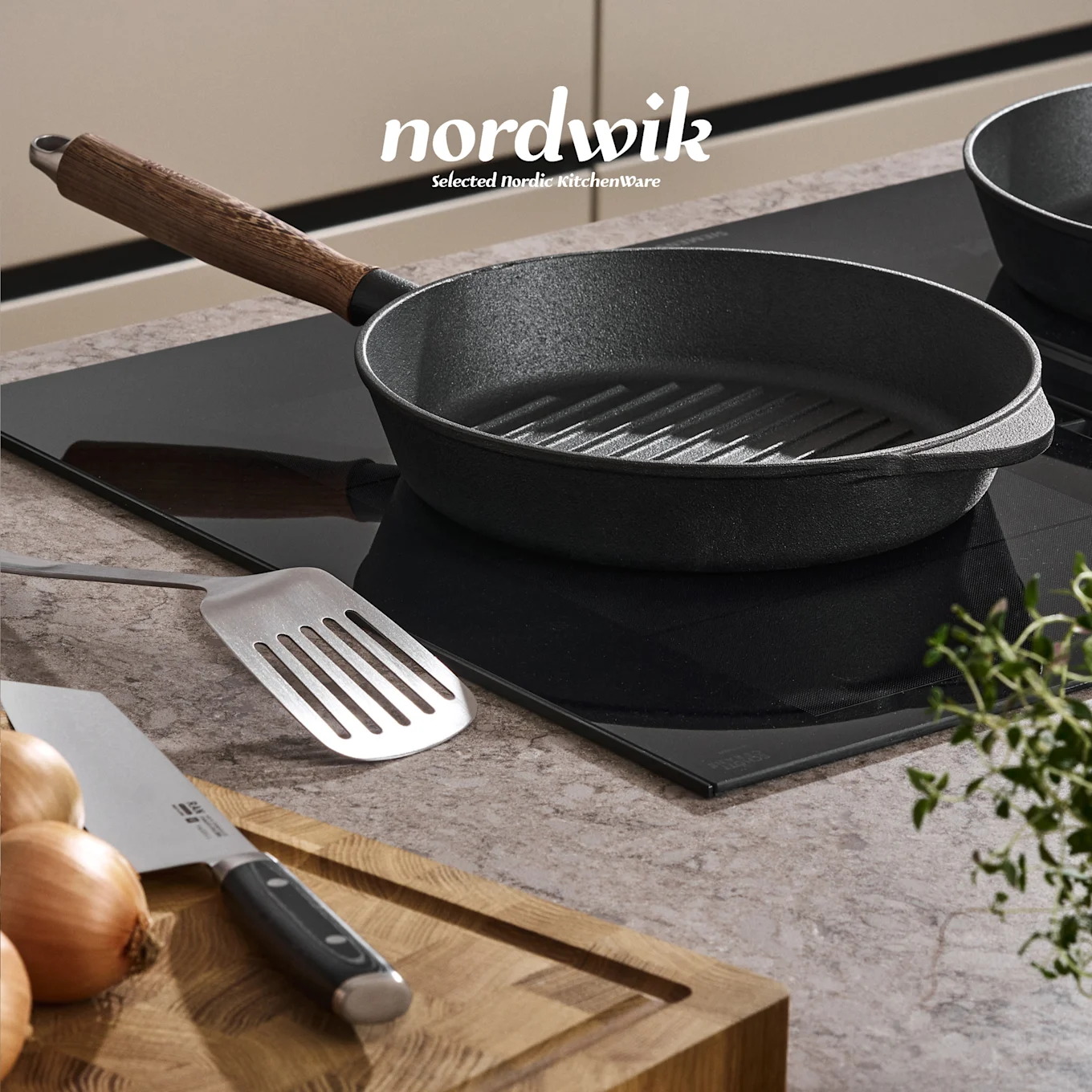 Musta valurautaparila induktioliedellä, lasta, sipuleita, veitsi ja timjamia puulaudalla. Nordwik-logo näkyvissä.