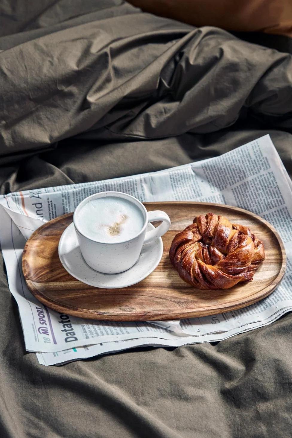 House Doctorin valko-harmaa Pion-kuppi cappuccinoa seisoo pienellä puualustalla, sanomalehden päällä, sängyllä, vieressä ruotsalainen kanelipulla.