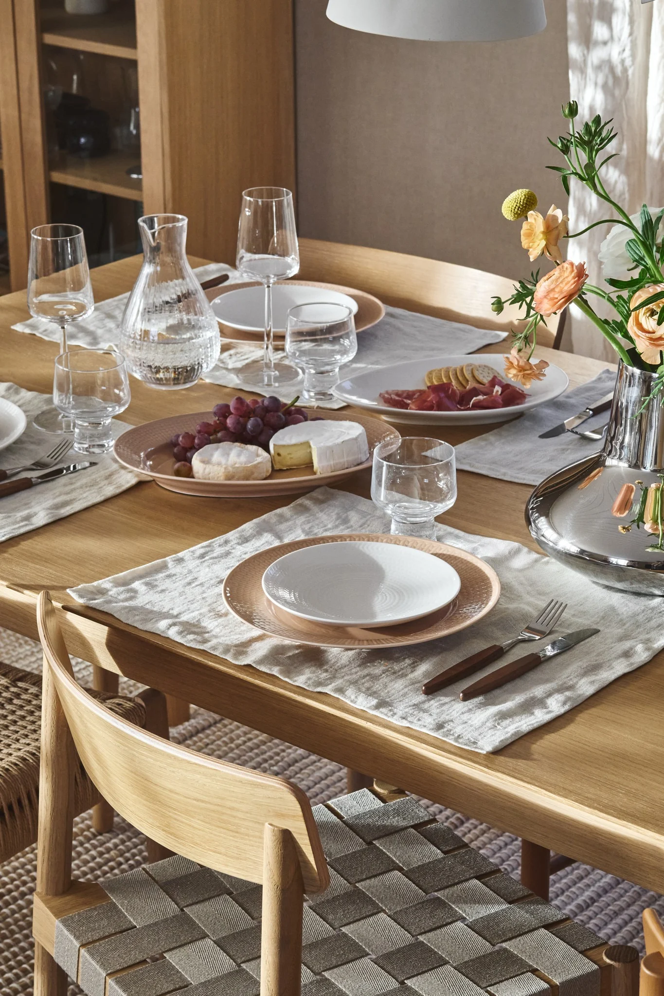 Puinen ruokapöytä astioilla, karahvilla, viinirypäleillä, juustoilla, leikkeleillä ja kukilla. Punotut tuolit.