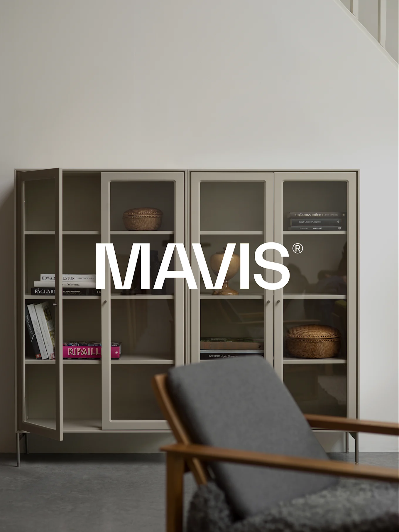 Kaksi vaaleanharmaata kaappia lasiovilla, joissa on kirjoja ja sisustustavaroita. Puinen tuoli on sumea etualalla. 'MAVIS®'-logo on keskitetty.
