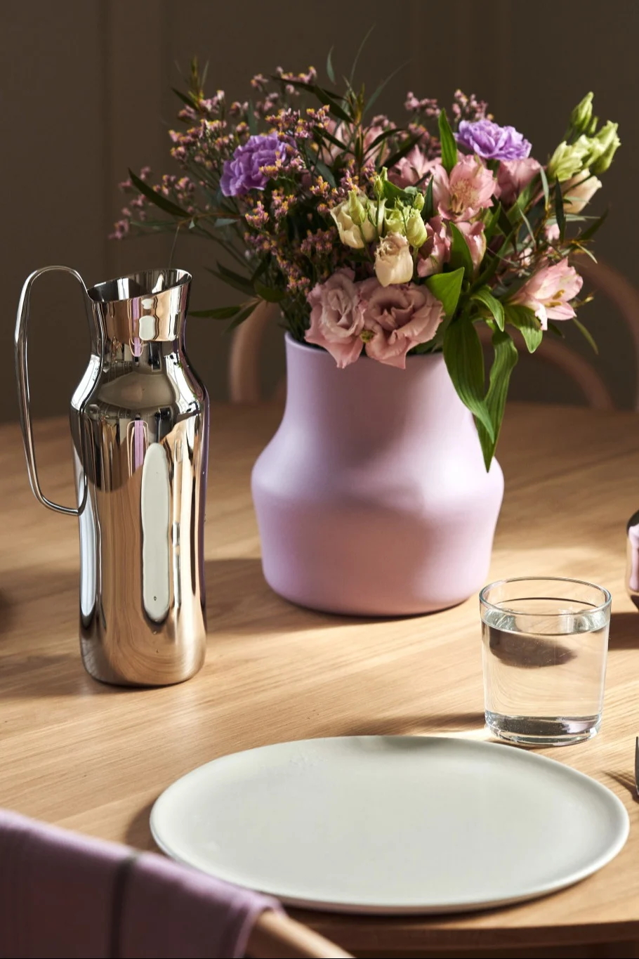 Tässä näet violetin Dorotea-maljakon, jossa on kimppu vaaleanpunaisia ja violetteja kukkia, seisomassa pöydällä hopeisen Dorotea-maljakon vieressä.