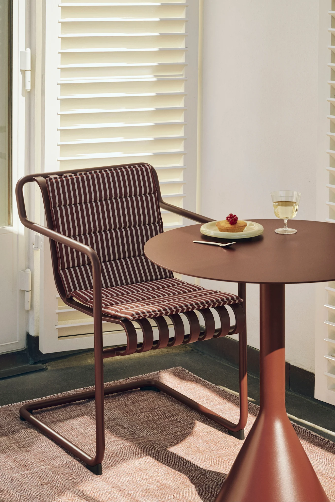 Punertavanruskea tuoli raidallisella tyynyllä yhteensopivan pyöreän pöydän äärellä, pöydällä kakkua ja valkoviiniä.