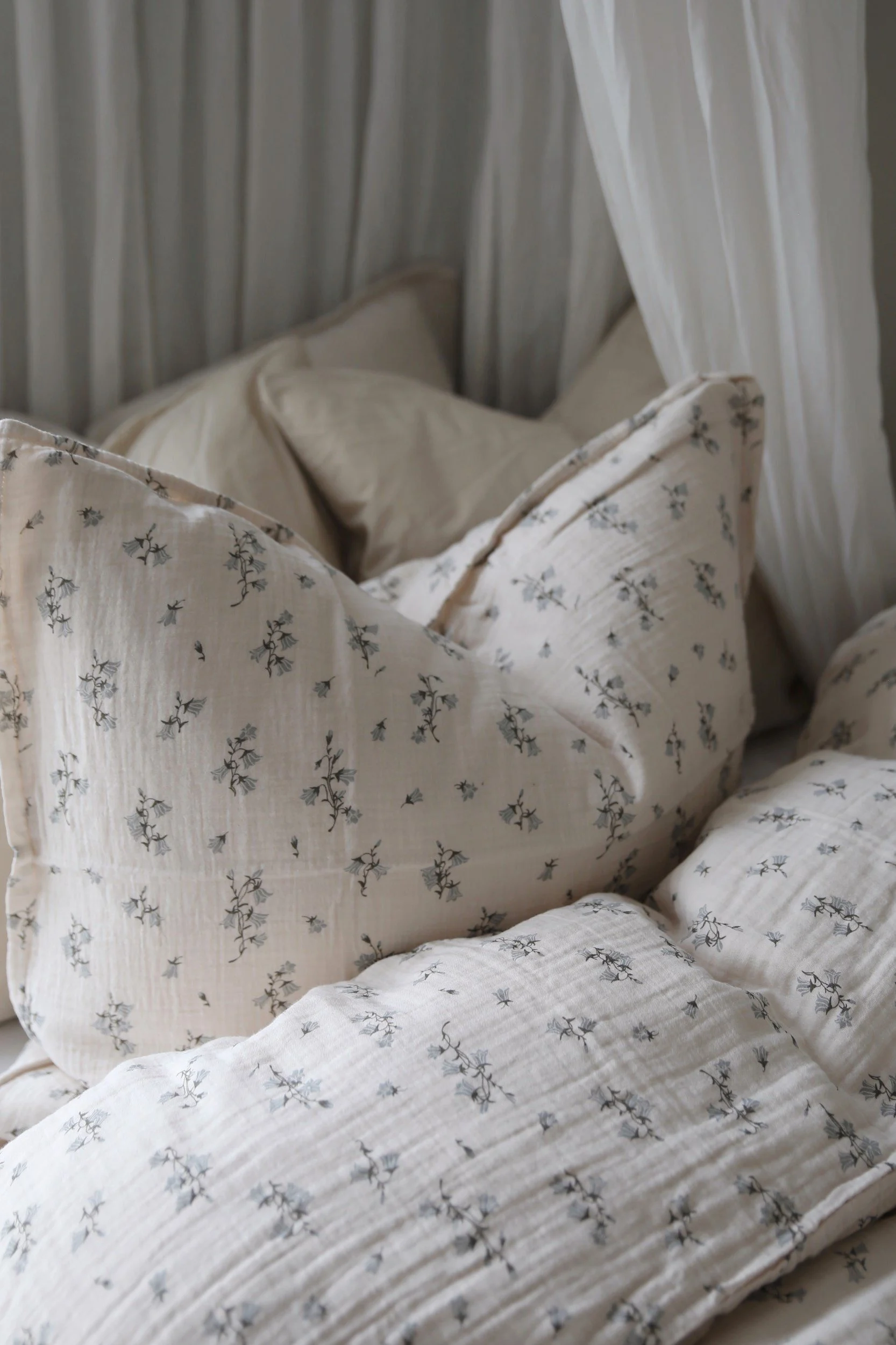 Kodikkaat vuodevaatteet vaaleasta, beigen sävyisestä kreppikankaasta, jossa on hienovarainen sininen kukkakuvio, sekä valkoiset verhot.