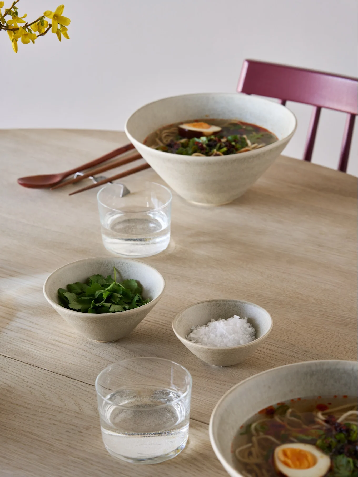 Kaksi ramen-kulhoa munien ja yrttien kera, lisukekulhoja korianteria ja suolaa, vettä, syömäpuikkoja ja lusikka vaalealla puupöydällä.