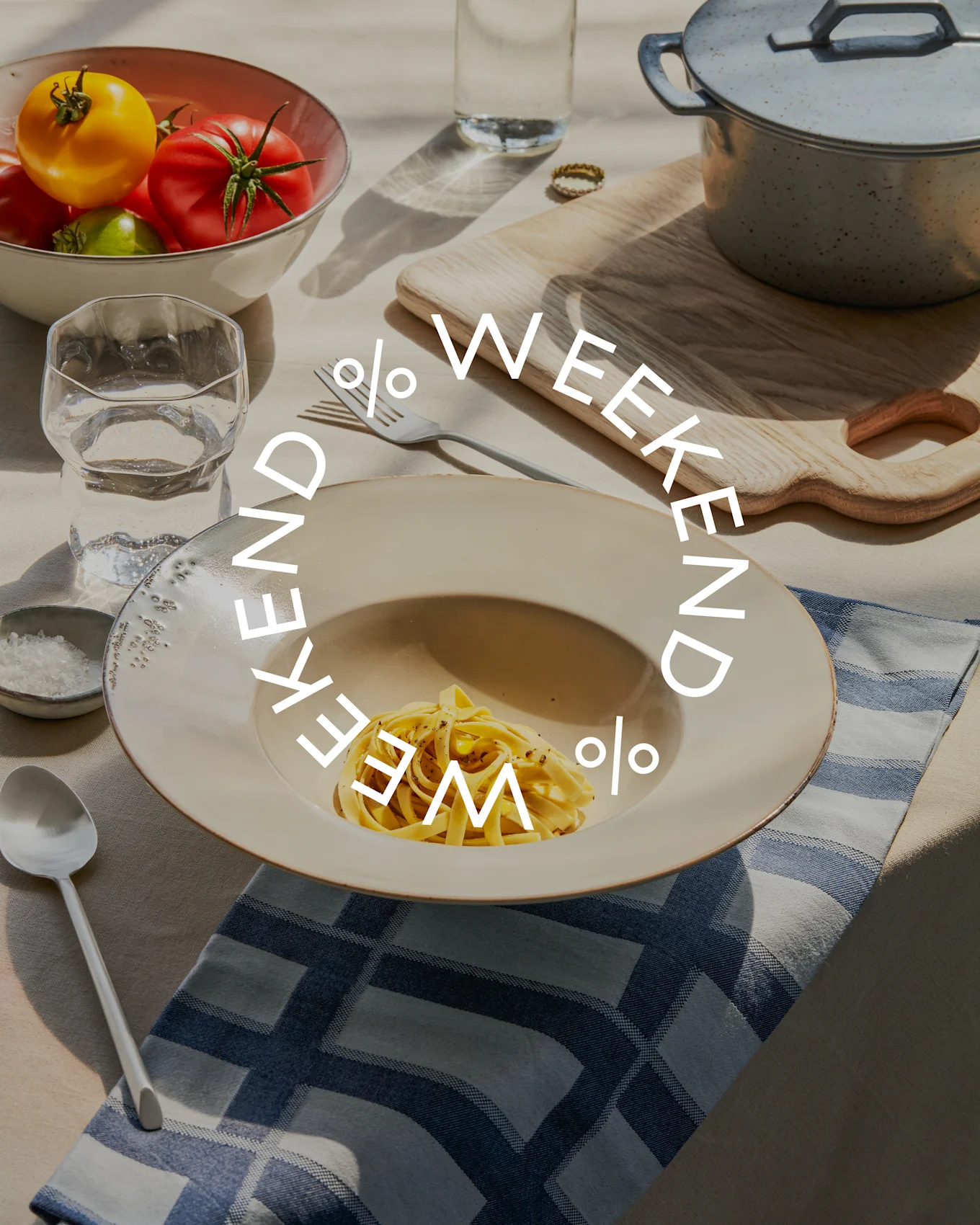 Pöytä katettuna pastalla, kulholla tuoreita tomaatteja, vesilasilla ja kattilalla. Teksti 'WEEKEND % WEEKEND %' on päällä.