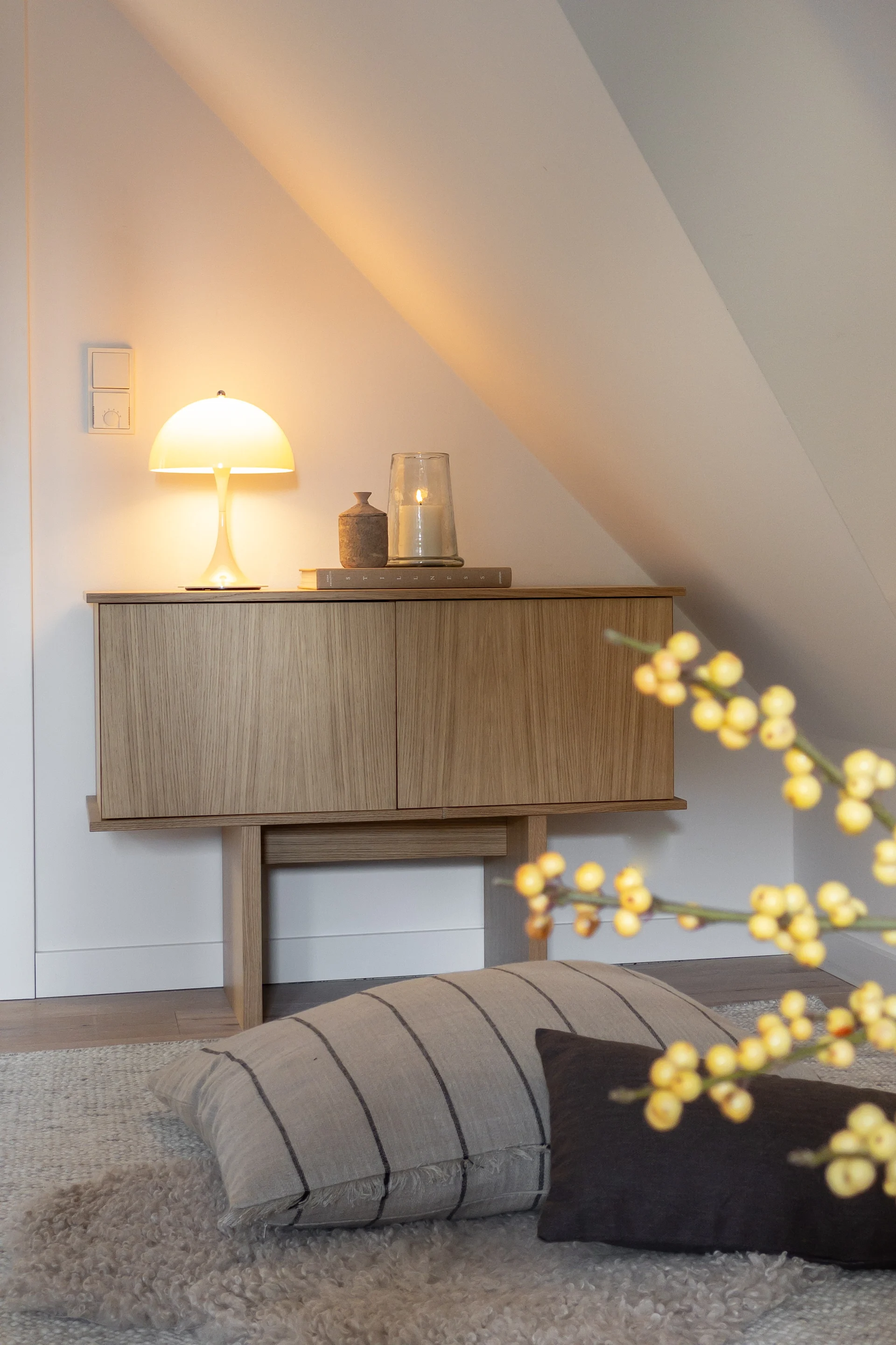 Puinen kaappi, jossa hehkuva sienilamppu ja palava kynttilä, valkoista, kaltevaa seinää vasten viihtyisässä huoneessa.