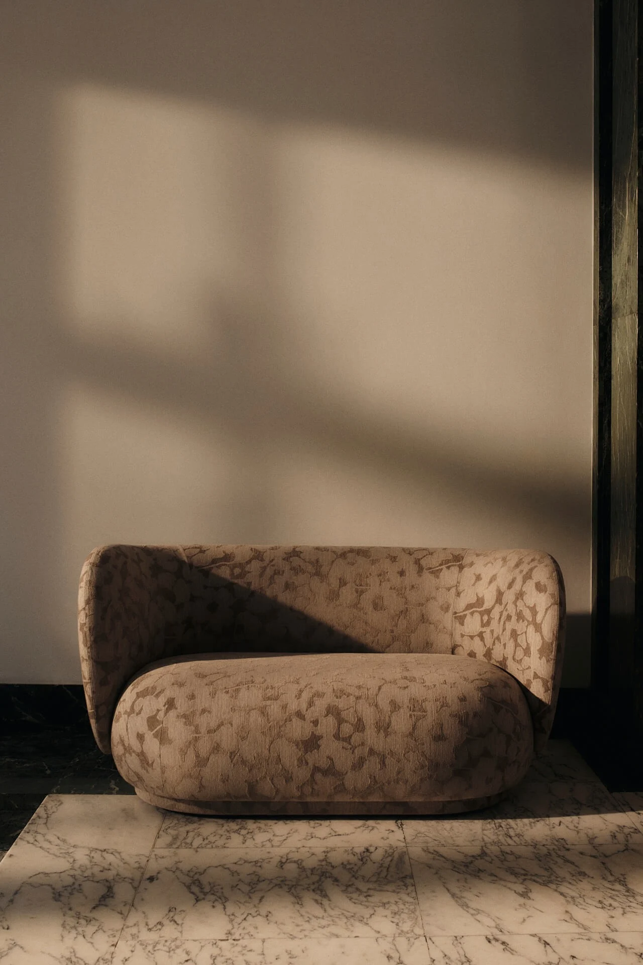 Pyöreä beige nojatuoli hienostuneella kuviokankaalla valkoisella marmorilattialla, lämpimän valon luomine dramaattisine varjoineen.