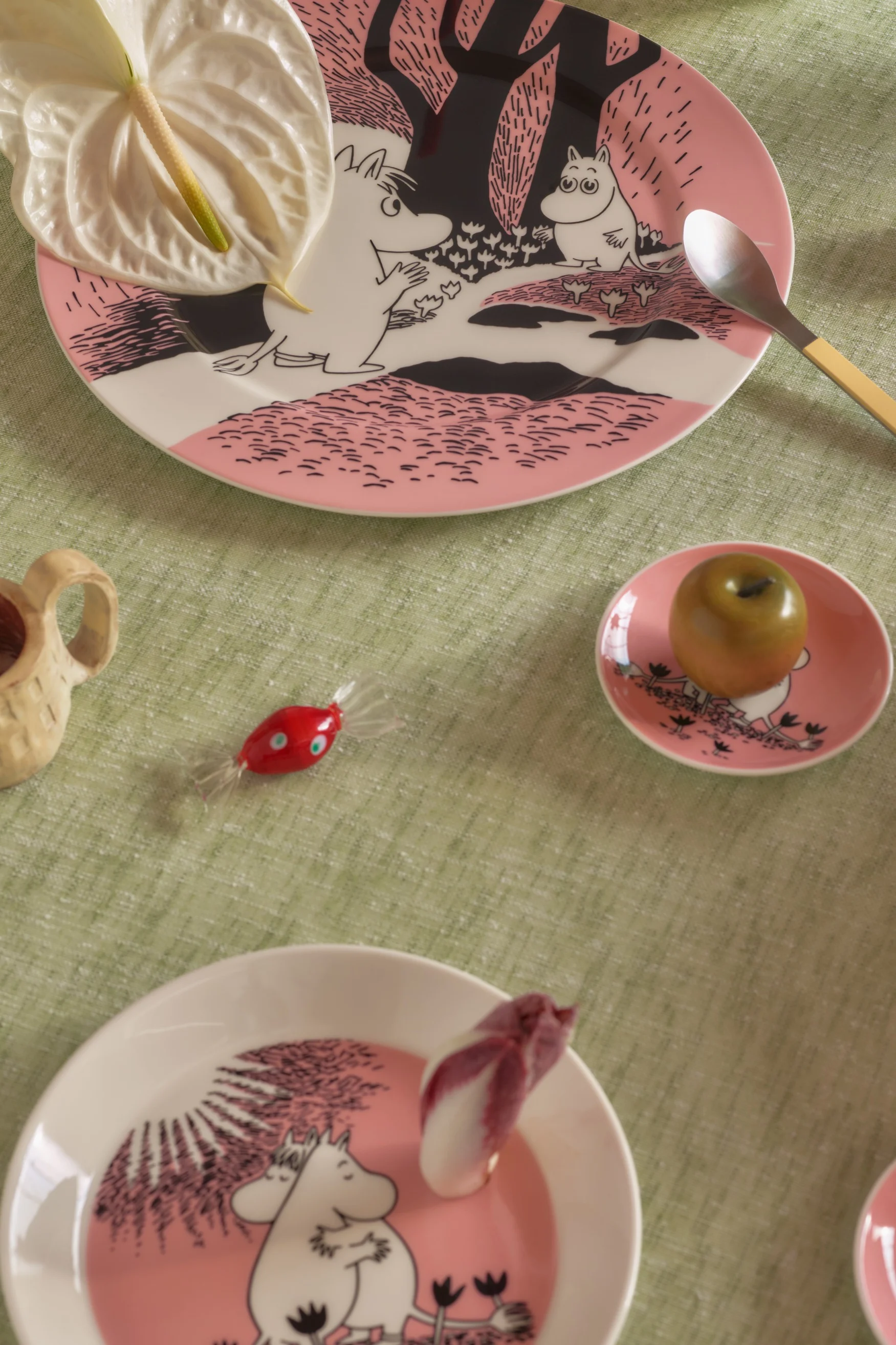 Pöytä katettu vaaleanpunaisilla Muumi-lautasilla, flamingonkukalla, lusikalla, vihreällä omenalla, punaisella karkilla ja puukupilla vihreällä pöytäliinalla.