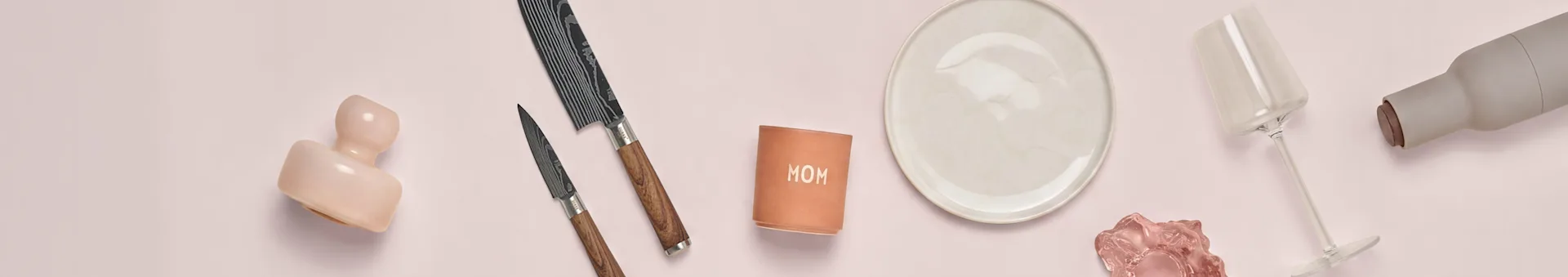 Pinkki keramiikka, veitset, 'MOM' muki, lautanen, pinkki lasi, viinilasi ja harmaa pullo pinkillä taustalla.
