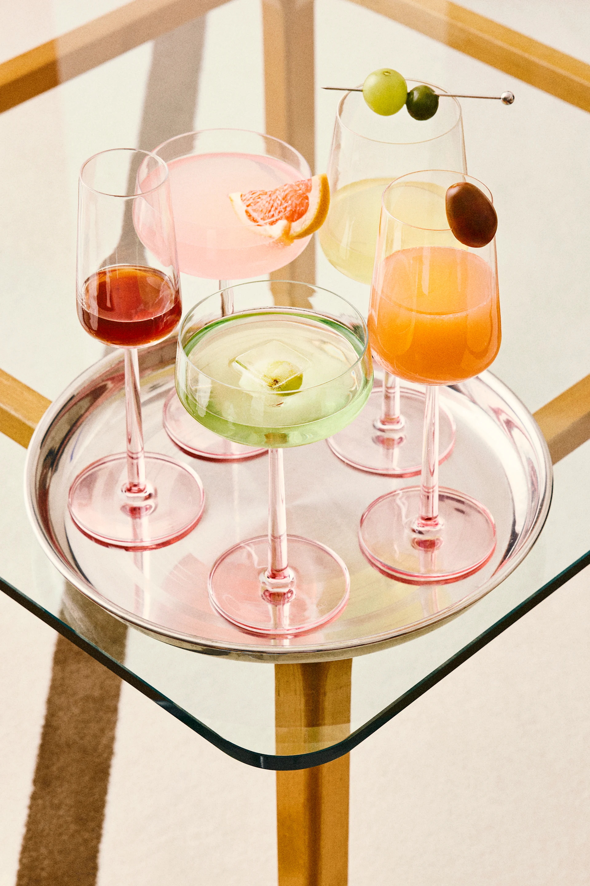 Viisi värikästä cocktailia hopeatarjottimella, mukaan lukien pinkkejä, vihreitä, oransseja ja punaisia juomia erilaisilla koristeilla, lasipöydällä.