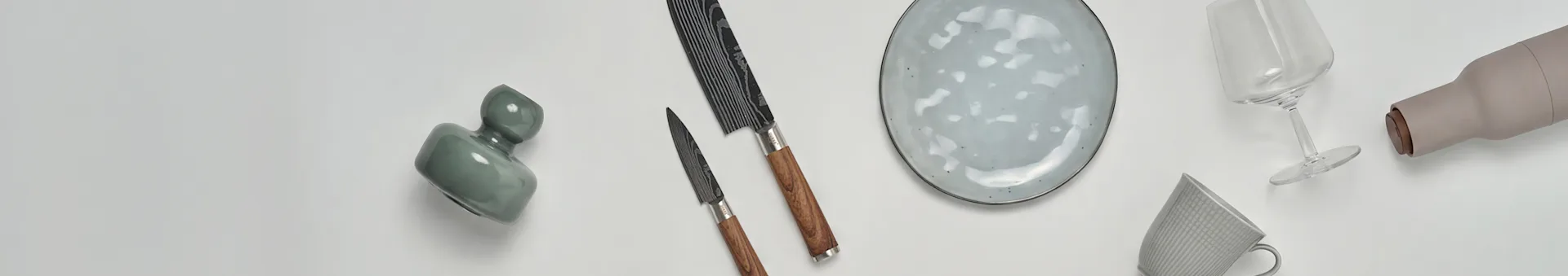 Erilaisia keittiö- ja ruokailuvälineitä, kuten vihreä maljakko, kaksi veistä, pilkullinen lautanen, viinilasi, beige pullo ja kuvioitu muki.