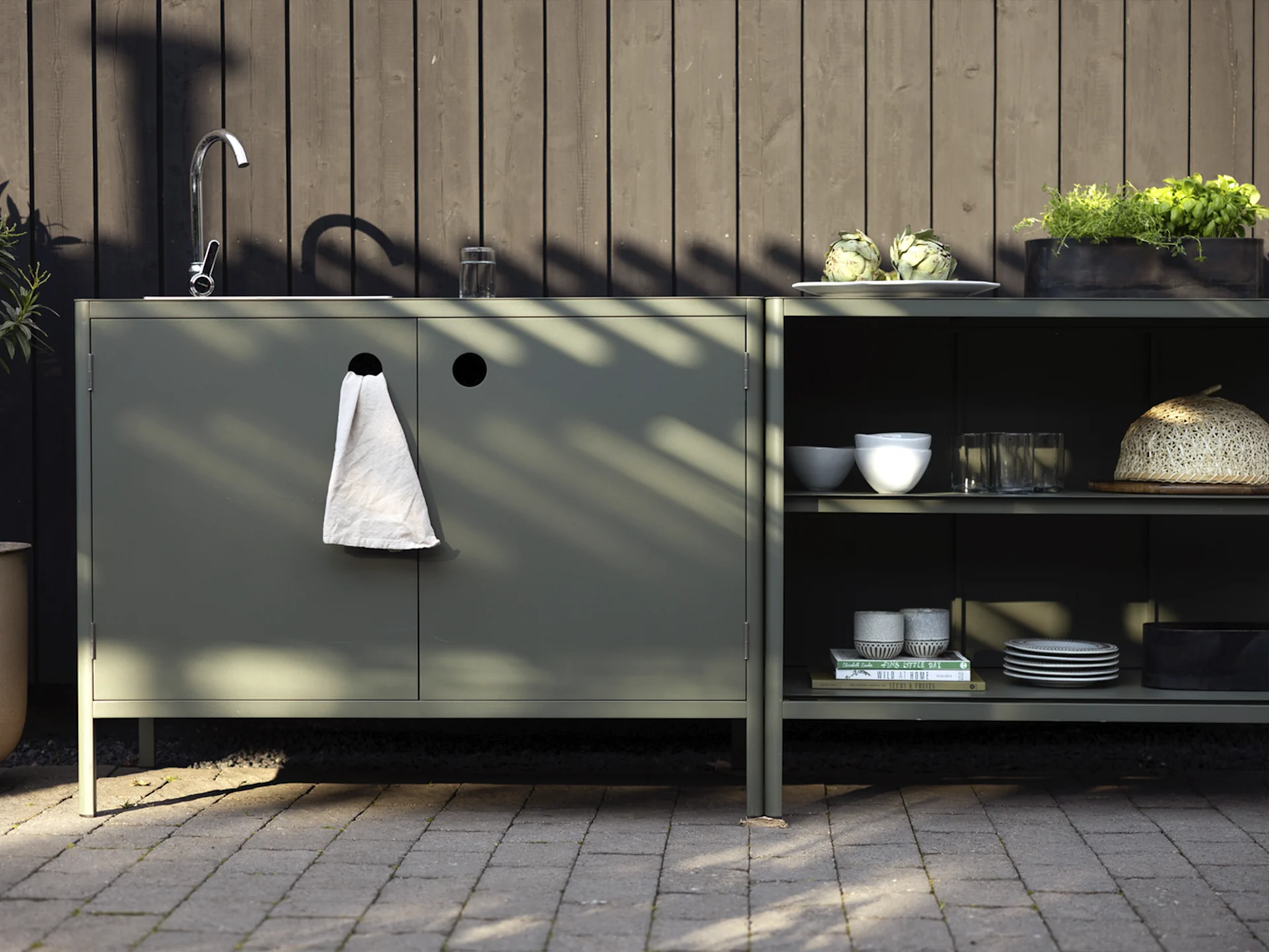 Ulkoikeittiö tummanvihreillä kaapeilla, altaalla, hanalla ja hyllyillä, joissa artisokkia, yrttejä ja astioita.