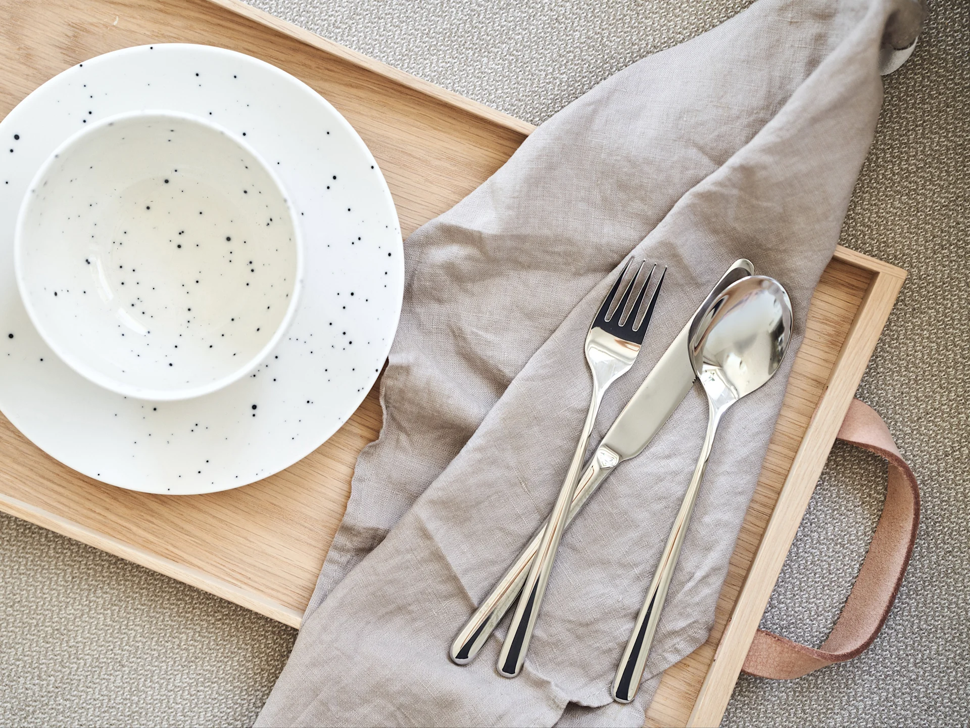Puinen tarjotin, jossa on valkoinen pilkullinen kulho ja lautanen, harmaa pellavaservetti ja hopeiset aterimet kudotulla matolla.