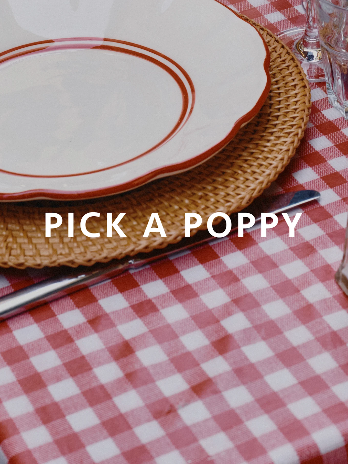 Katettu pöytä, punavalkoruudullinen pöytäliina, pajualusta ja valkoinen lautanen punaisella reunalla. Teksti: PICK A POPPY.