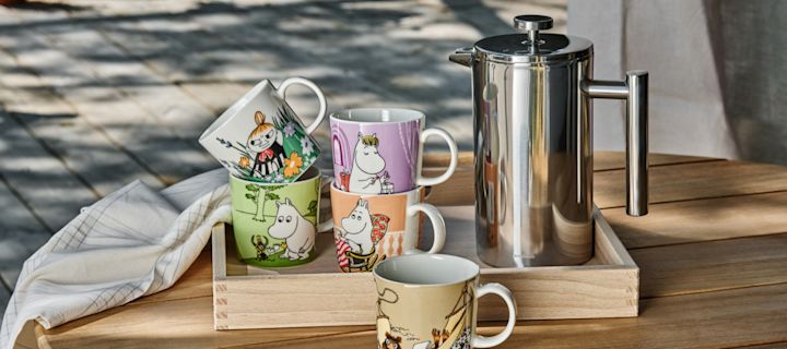 Moomin Arabian erilaisia Muumi-kuppeja seisoo pöydällä ulkona, leimasinkannun vieressä.