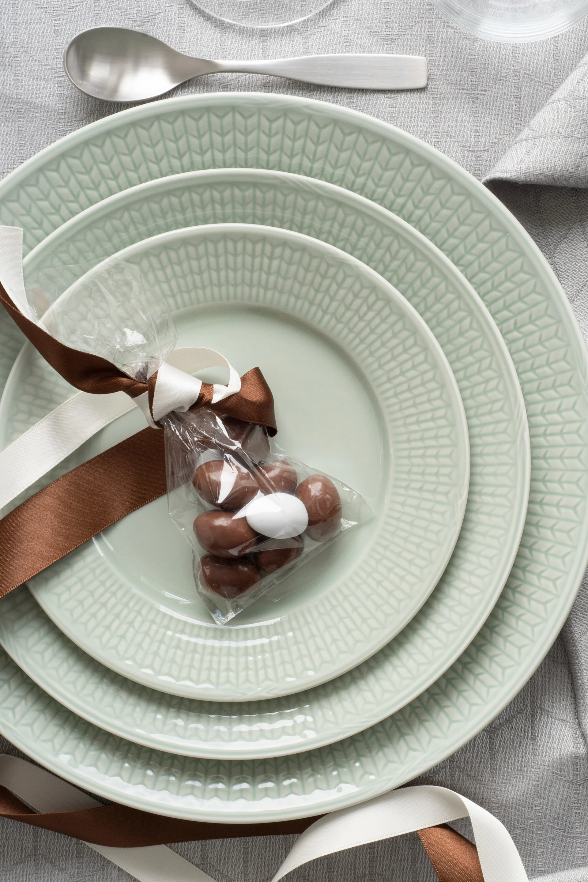 Pinottuja vaaleanvihreitä lautasia, lahjapussillinen praliineja harmaalla pöytäliinalla, hopeisten aterimien vieressä.