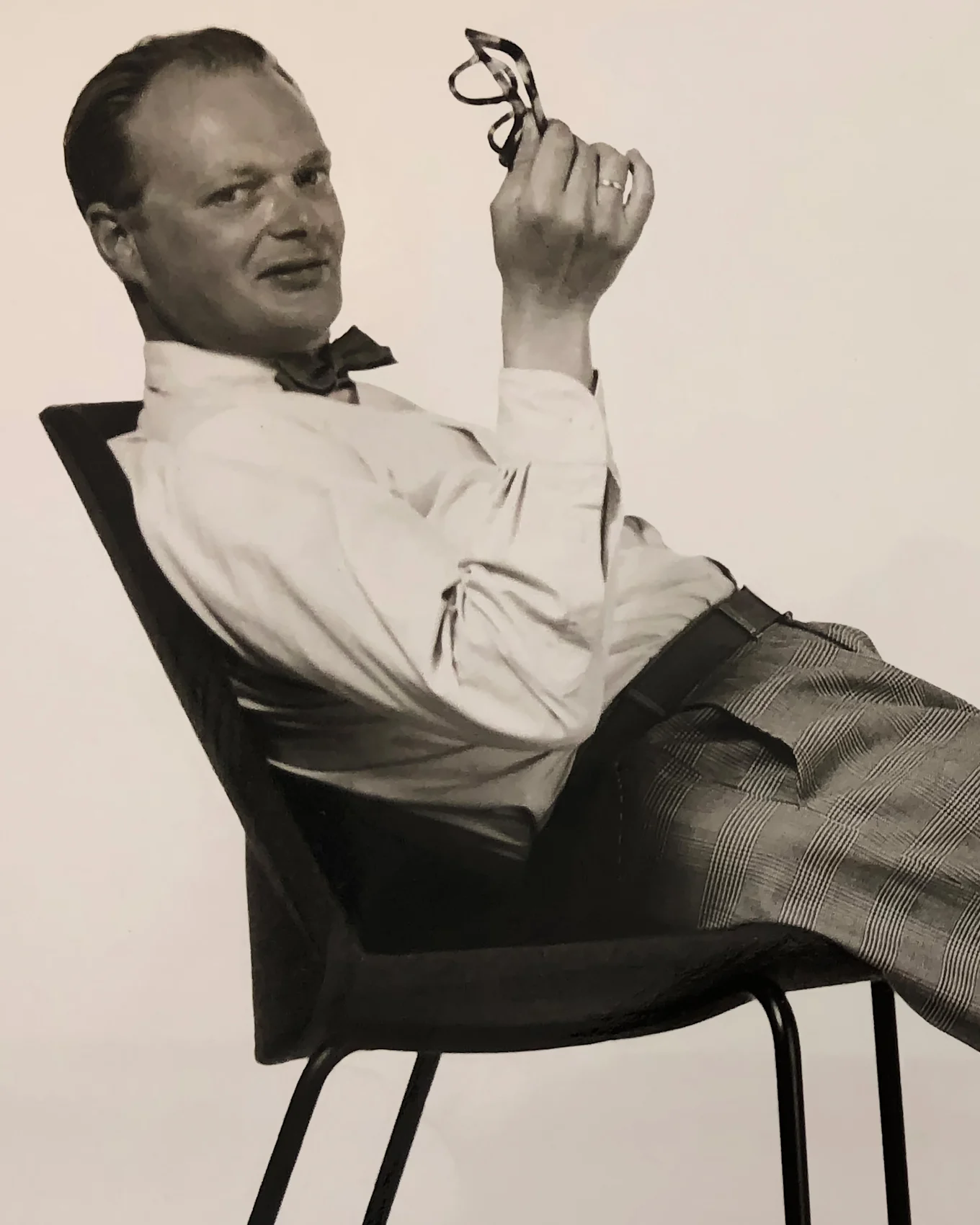 Mies rusetissa ja ruudullisissa housuissa nojaa tuolissa, pitää silmälaseja ja hymyilee kameralle.
