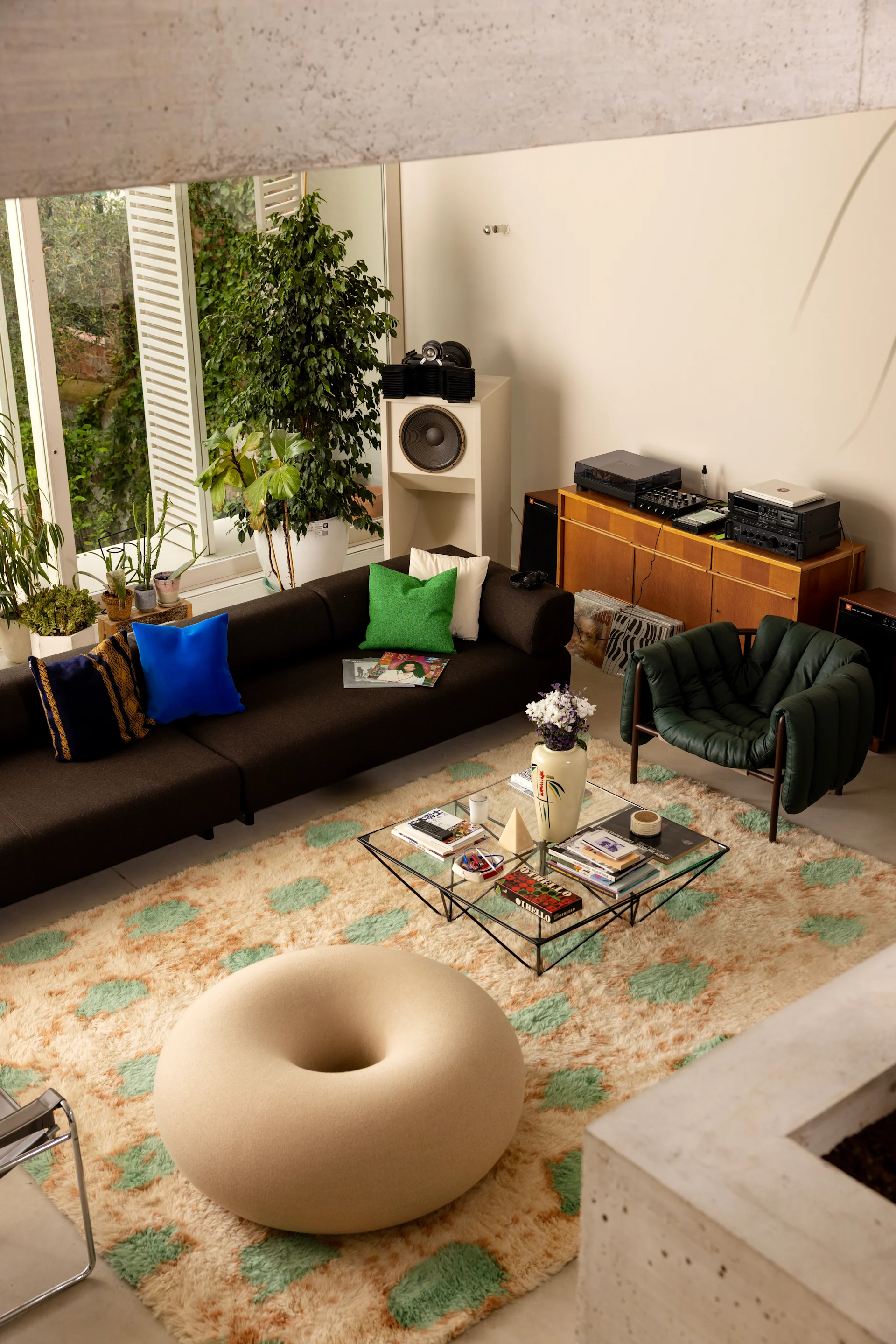 Donitsinmuotoinen, värikäs puff, joka on sijoitettu minimalistiseen huoneeseen, jossa on suuret ikkunat, vangitsee vuoden 2026 sisustustrendien ytimen ja toimii leikkisänä mutta hienostuneena katseenvangitsijana.