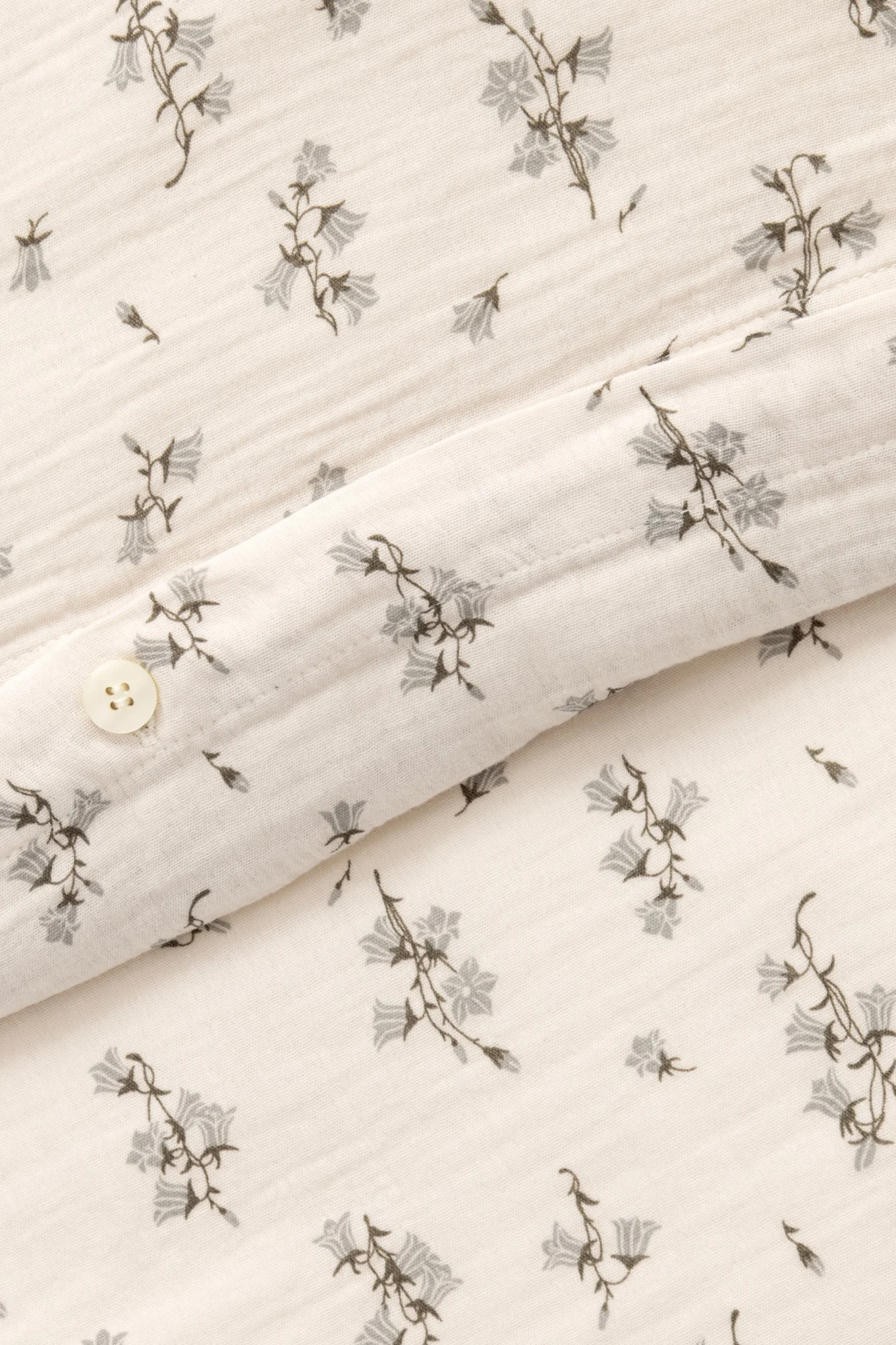 Lähikuva kermanvärisestä kankaasta, jossa on harmaa kellokukkakuvio ja valkoinen nappi.