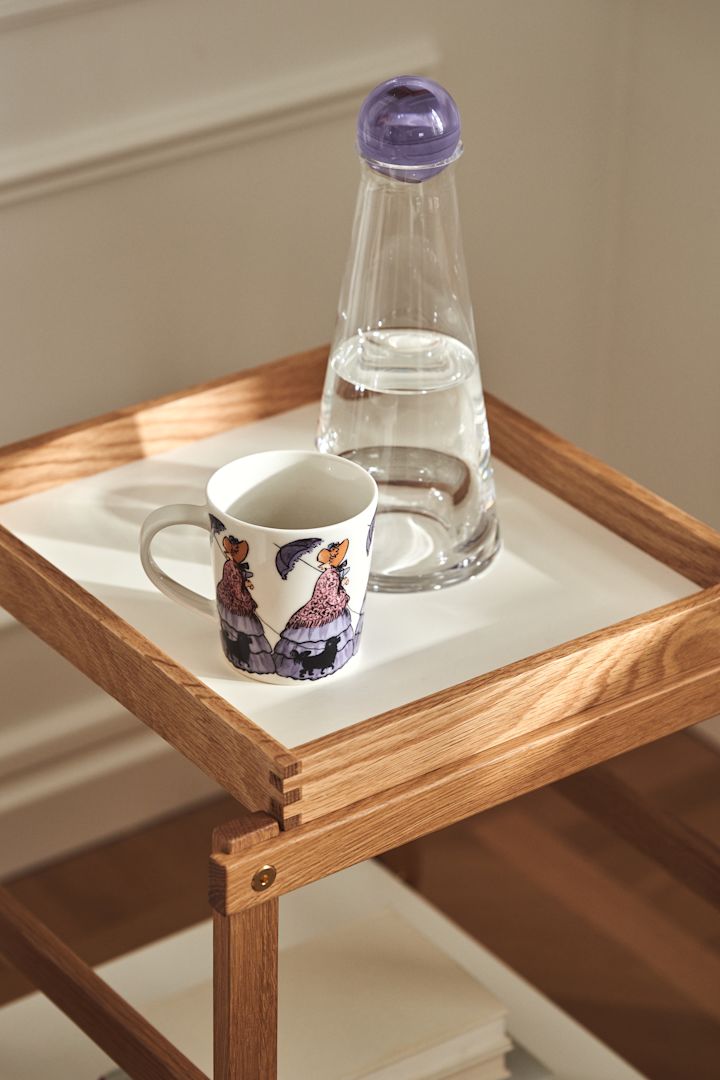 Muki, jossa on Elsa Beskow -motiivi Aunt Gredelin Design House Stockholmilta, ja Fia-karahvi lasista, jossa on violetti kansi, Frame-hylly sävyssä tammi ja valkoinen.