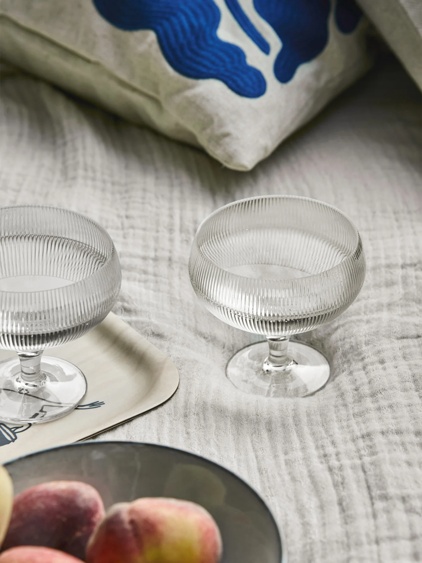 Kaksi uurrettua lasijälkiruokakulhoa, tyyny sinisellä abstraktilla taiteella, puutarjotin ja persikoita vaalealla kankaalla.