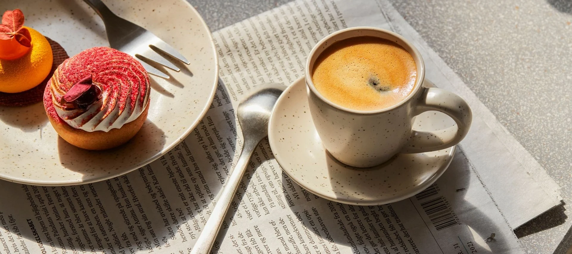 Kahvikuppi ja kaksi leivonnaista, punainen torttu ja oranssi, sanomalehden päällä.