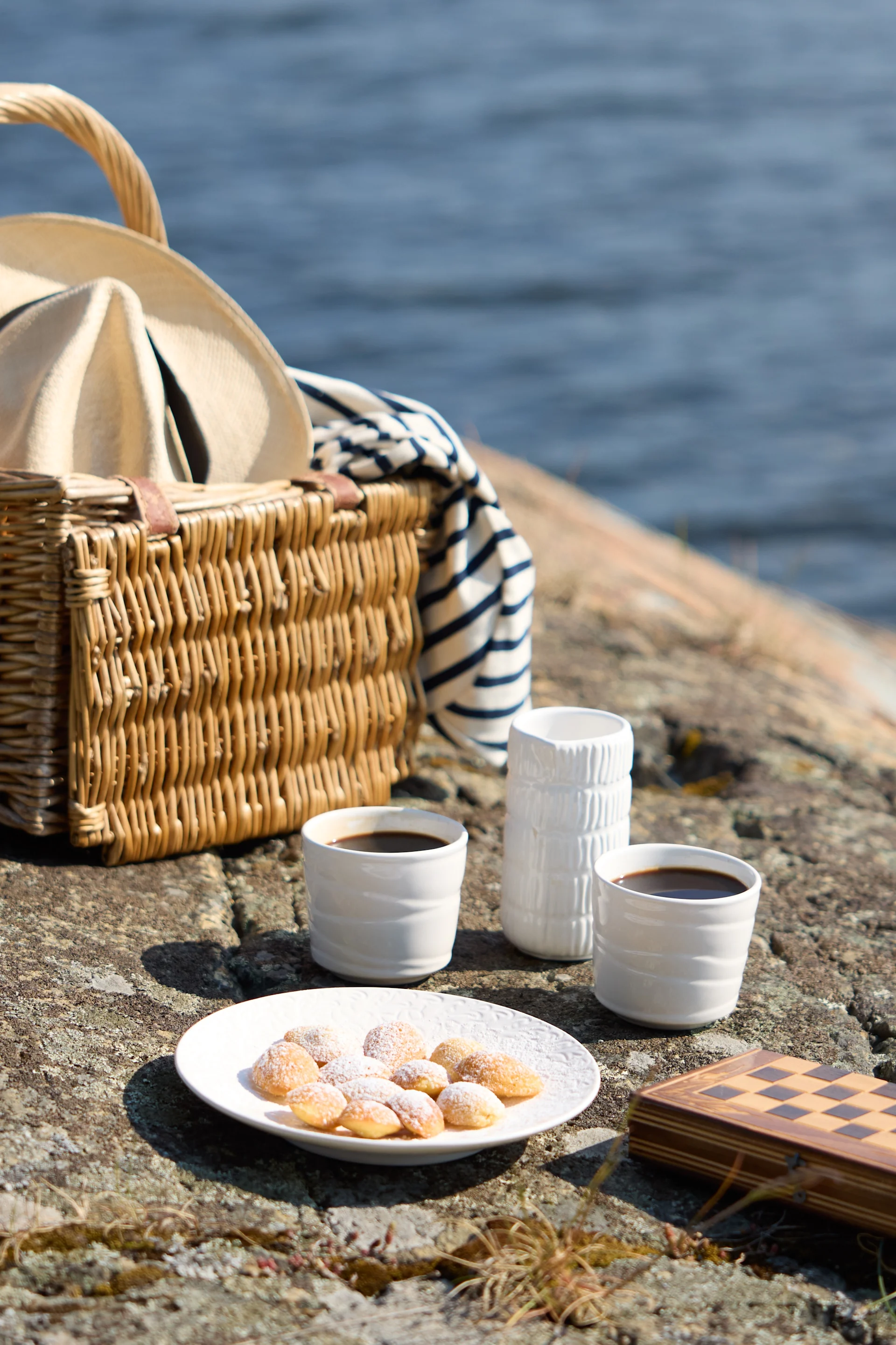 Piknik-kattaus kivikkoisessa maastossa veden äärellä, sisältäen kahvia, leivonnaisia ja shakkisetin.