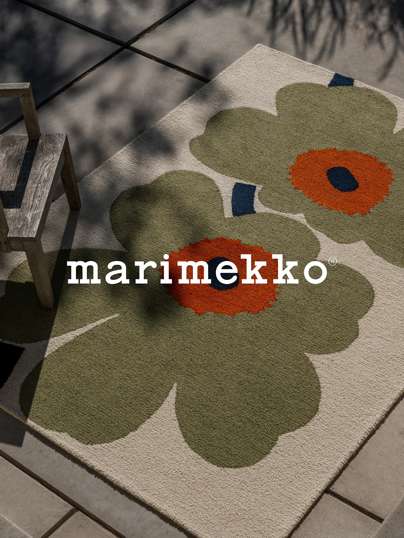 Marimekko-matto, jossa iso vihreä-oranssi kukkakuvio terassilla puutuolin vieressä.