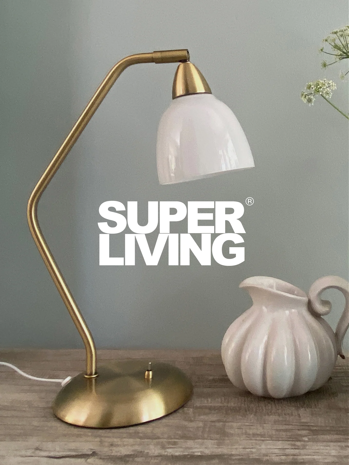 SUPER LIVING -brändi messinkisen pöytävalaisimen päällä, jossa valkoinen varjostin ja valkoinen, uurrettu kannu puupöydällä.